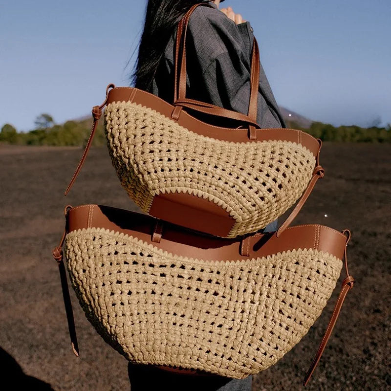 Two woven straw half-moon shoulder bags with brown leather trim and straps, shown being carried by a person outdoors in a dark field under a blue sky.