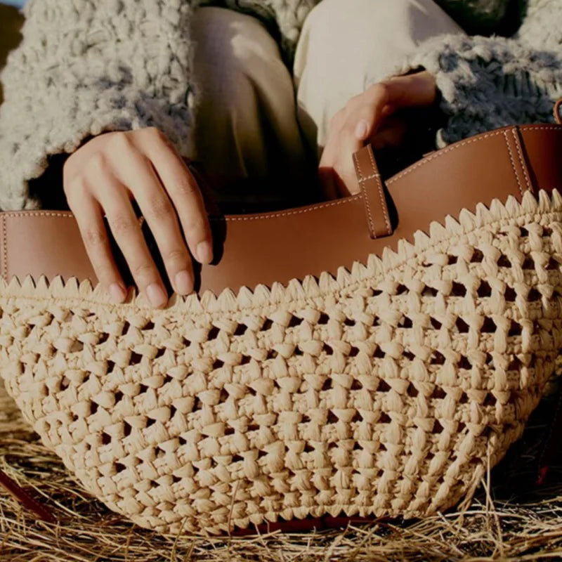 A close-up of a woven straw shoulder bag with a brown leather trim, held by a person wearing a textured gray sweater and beige pants on a straw-covered surface.