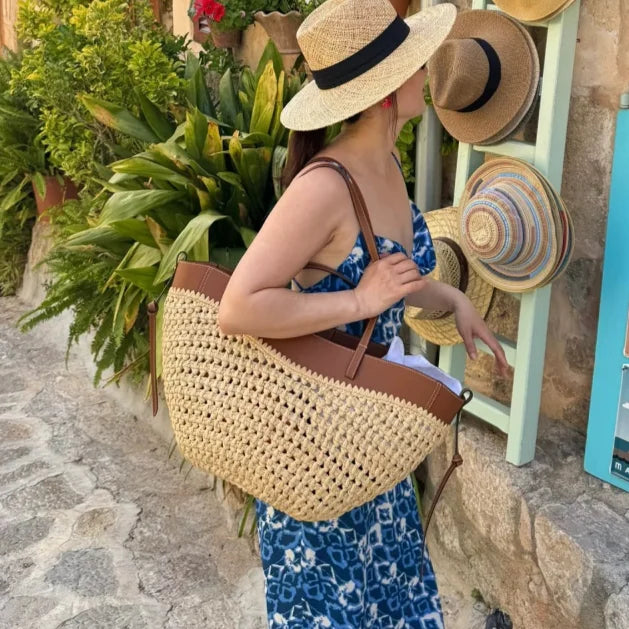 A woman wearing a straw hat and blue patterned dress carries a large woven straw tote bag with brown leather handles in an outdoor shop setting.