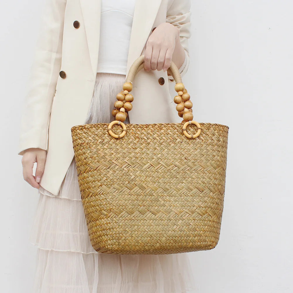 Woven straw tote bag with wooden handles held by a person wearing a white outfit on a light background