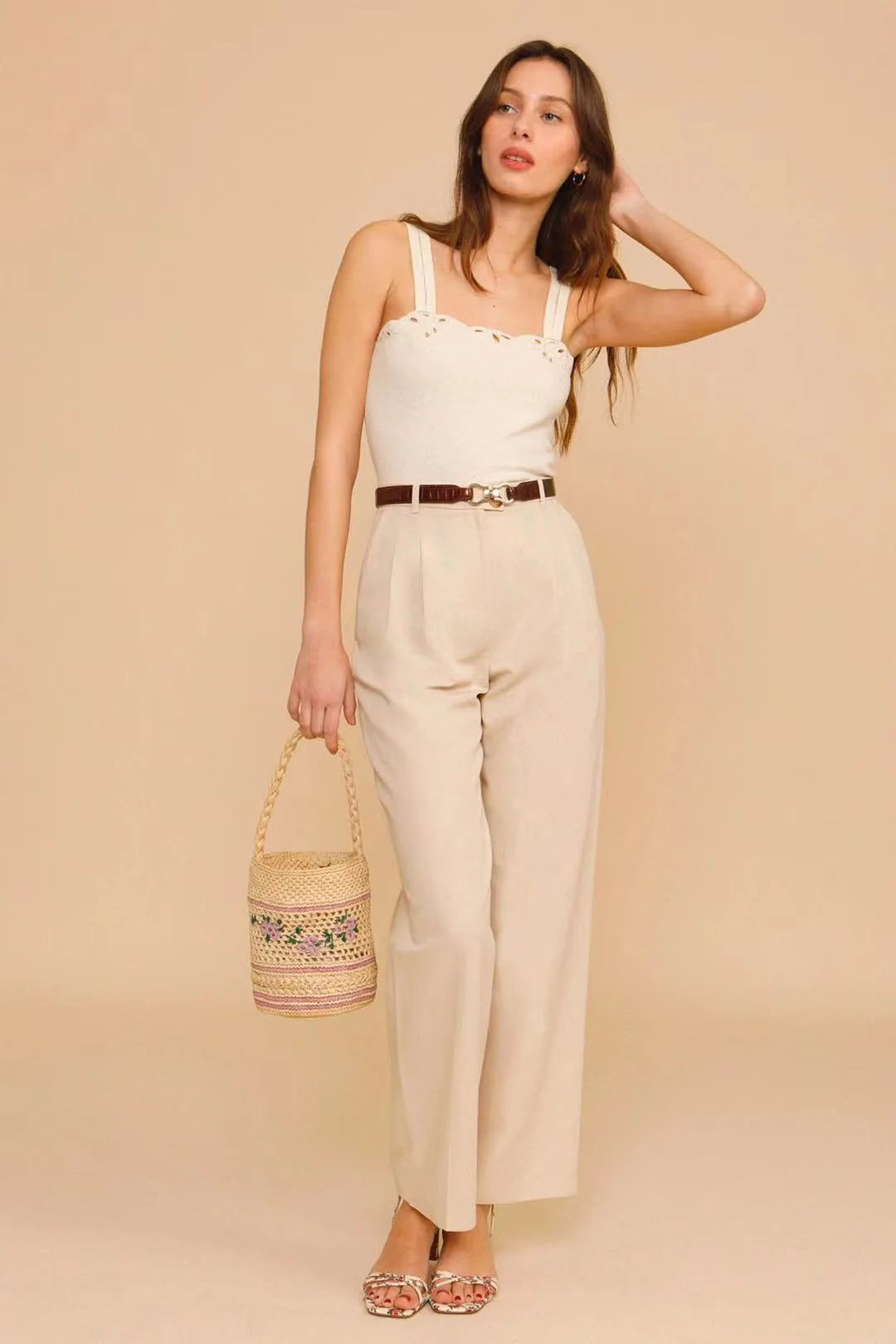 Woman wearing a beige jumpsuit holding a woven bag against a beige background