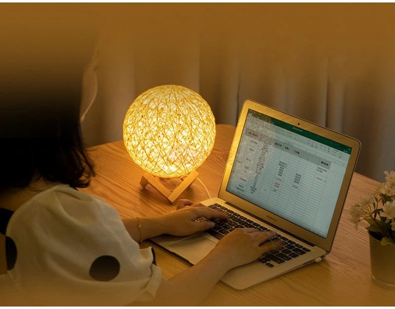 Person using a laptop with a glowing spherical lamp on a wooden desk.