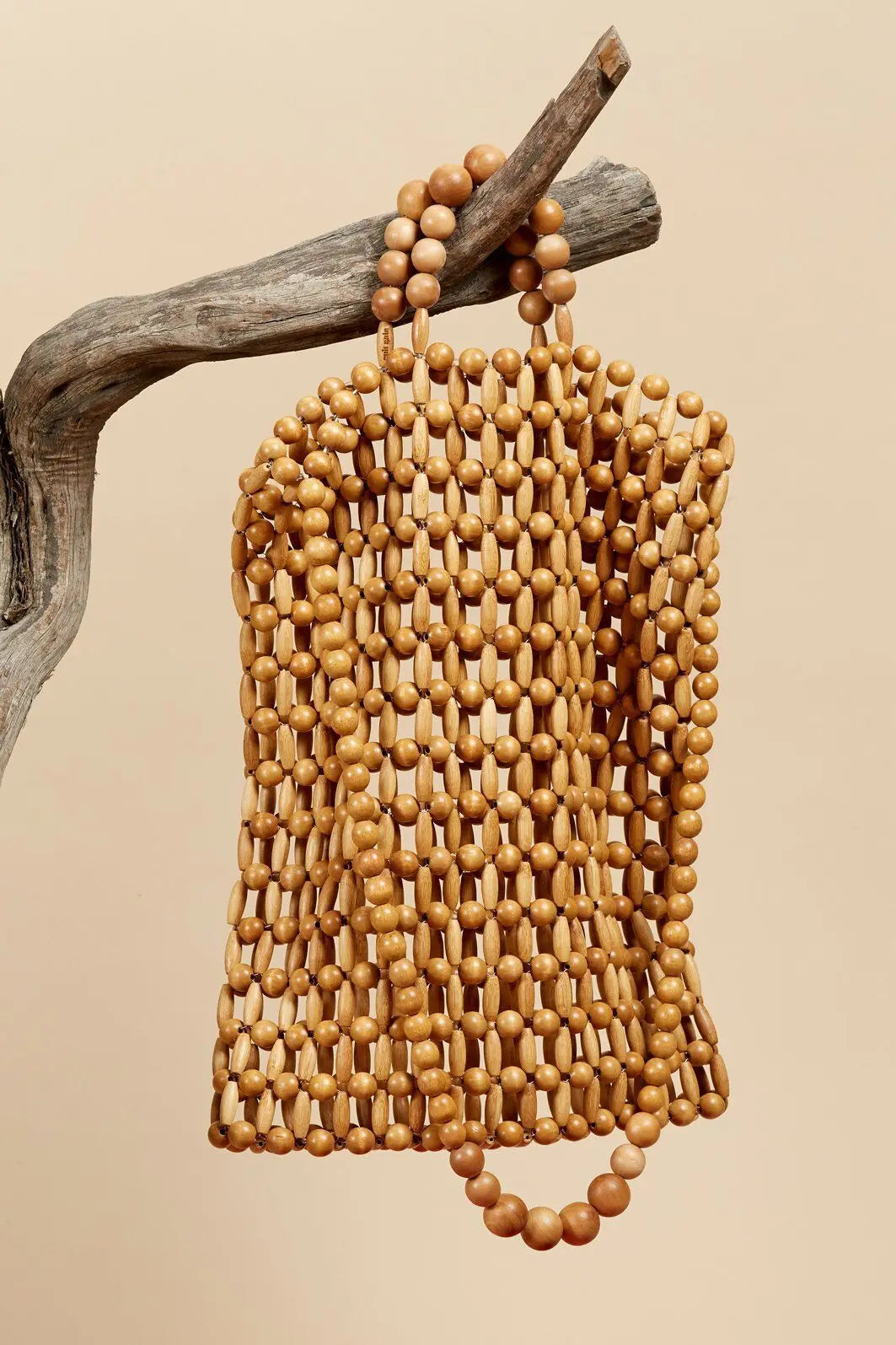 A natural wood beaded bag hanging from a rustic branch against a solid beige background.