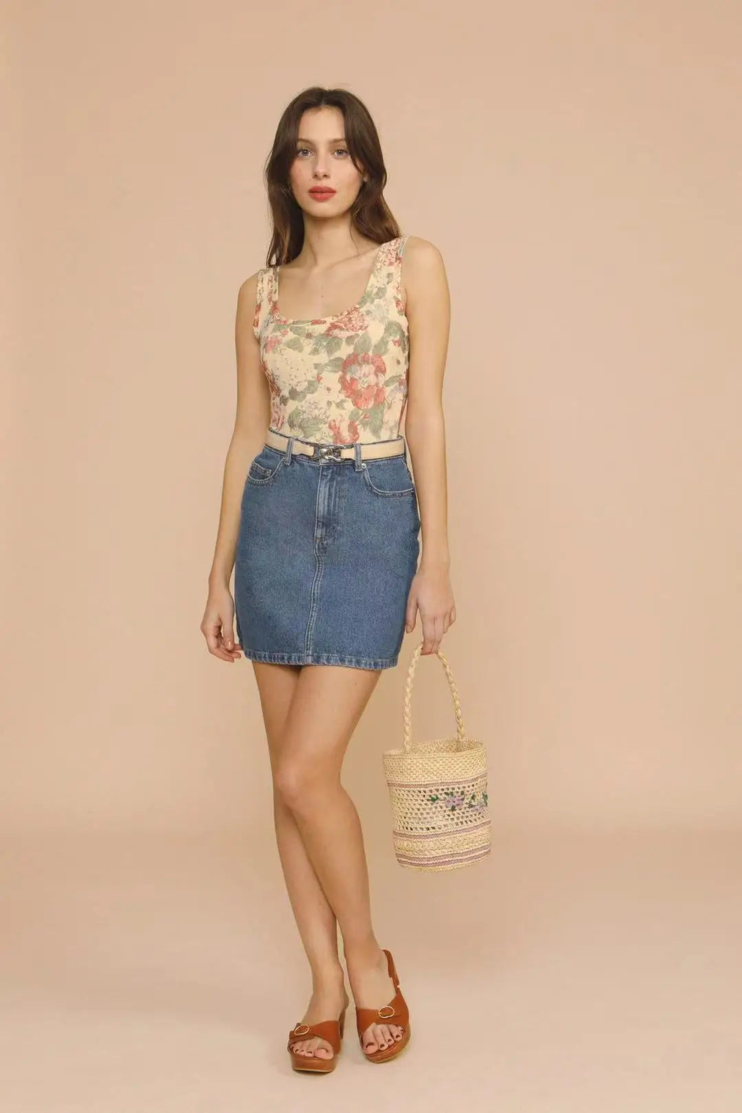 Woman wearing a floral top and denim skirt holding a yellow bag against a beige background