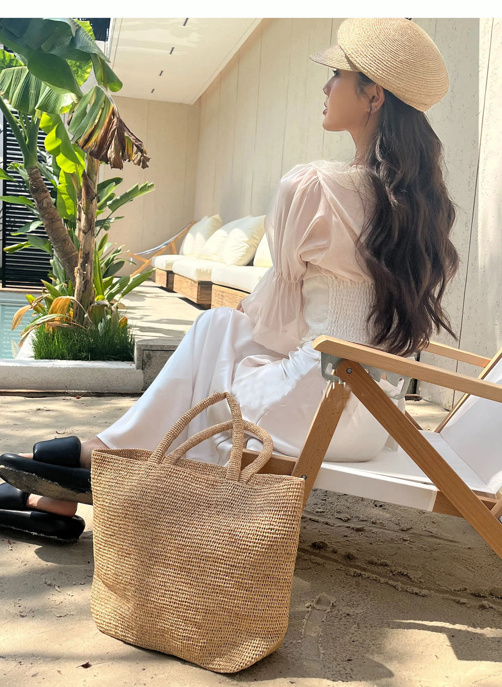 Woman sitting on a chair with a straw bag, wearing a hat and white outfit, in a sunlit outdoor setting.