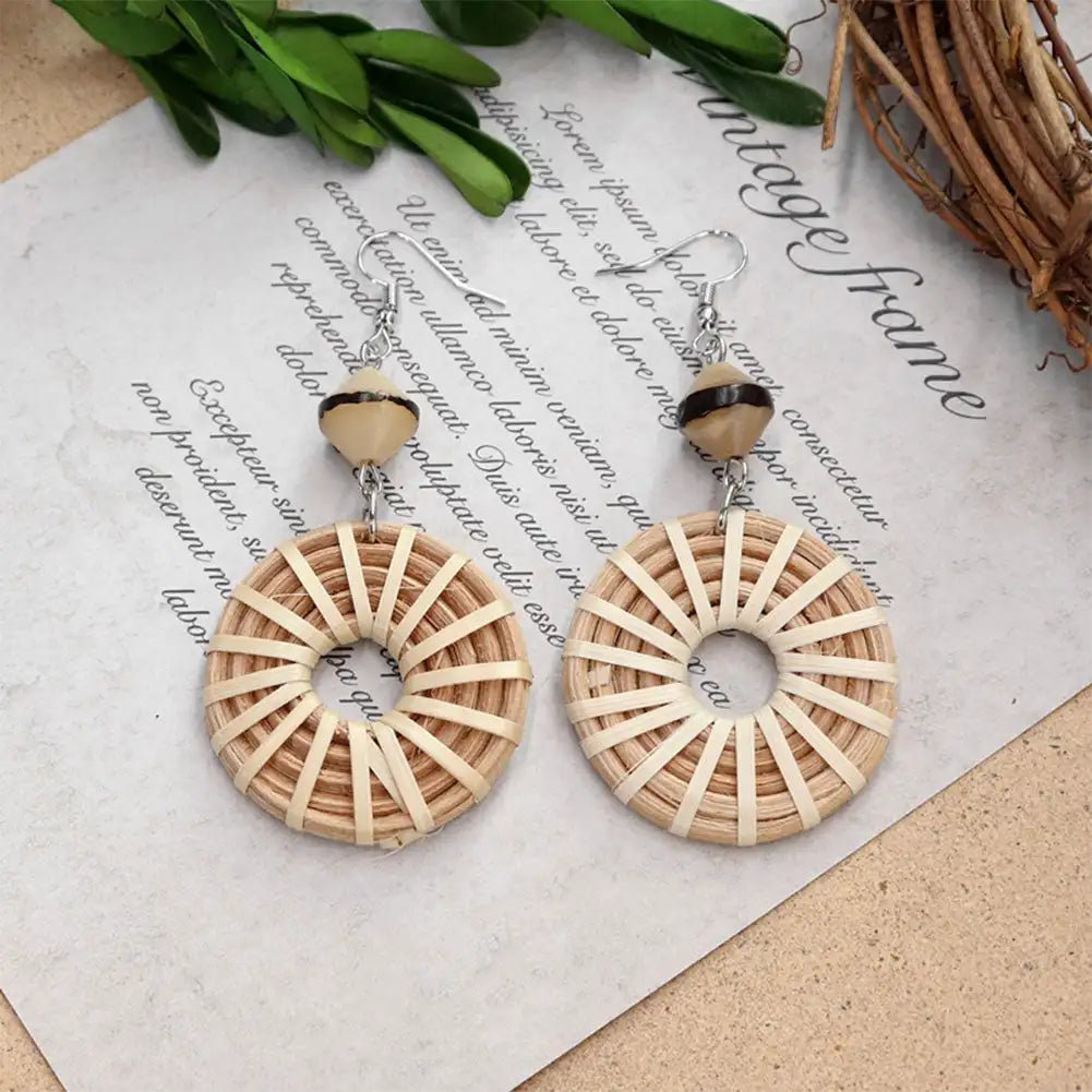 A pair of bohemian-style dangle earrings with circular woven rattan or straw disc charms and small dark beads, displayed on a piece of paper with cursive writing and decorative green leaves and twigs.