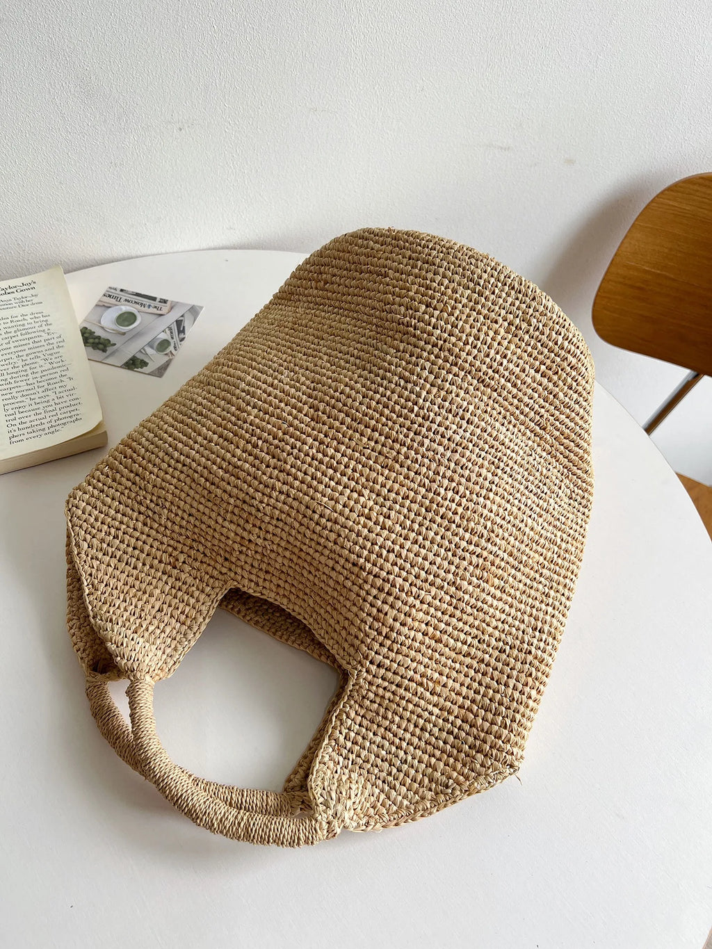 Woven beige bag on a white surface with a book and wooden chair in the background