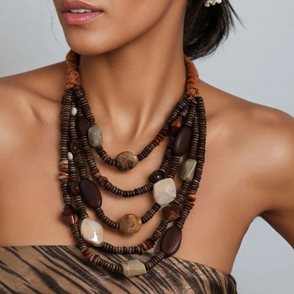 A close-up of a model wearing a multi-strand statement necklace with dark brown beads, natural stone accents, and a brown braided cord detail, paired with a brown and black patterned strap top.