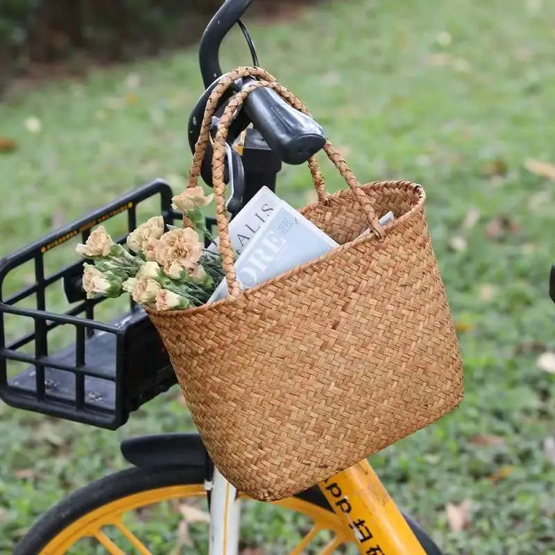 Woven basket attached to a bicycle with flowers and a book inside, on a grassy background