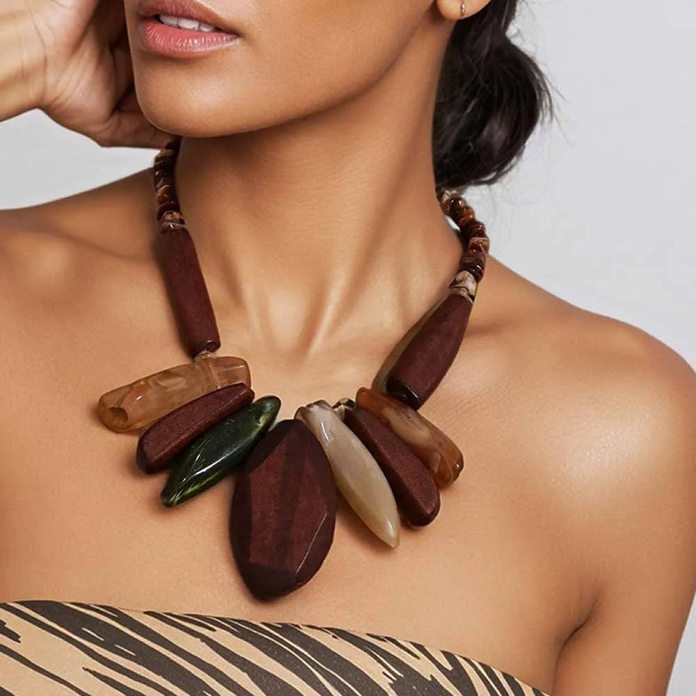 A close-up of a woman wearing a chunky statement necklace made of polished dark brown and earthy stone-like beads with a beige and green accent, paired with a beige and black patterned strap top.