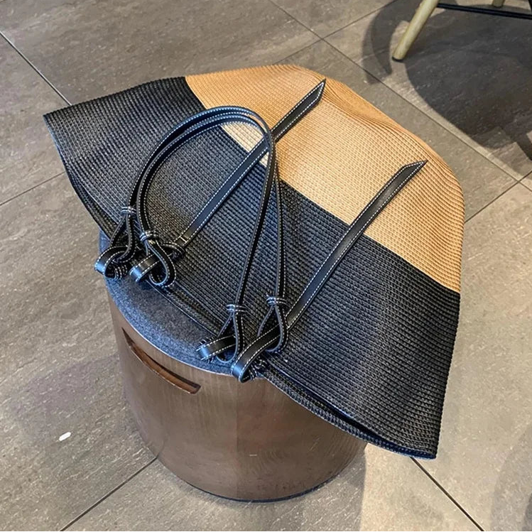 A woven tote bag with a color-blocked design in black and tan, resting on a round wooden stool in an indoor setting. The bag has black leather straps and is shown with the YUE JABON brand name visible in the top left corner.