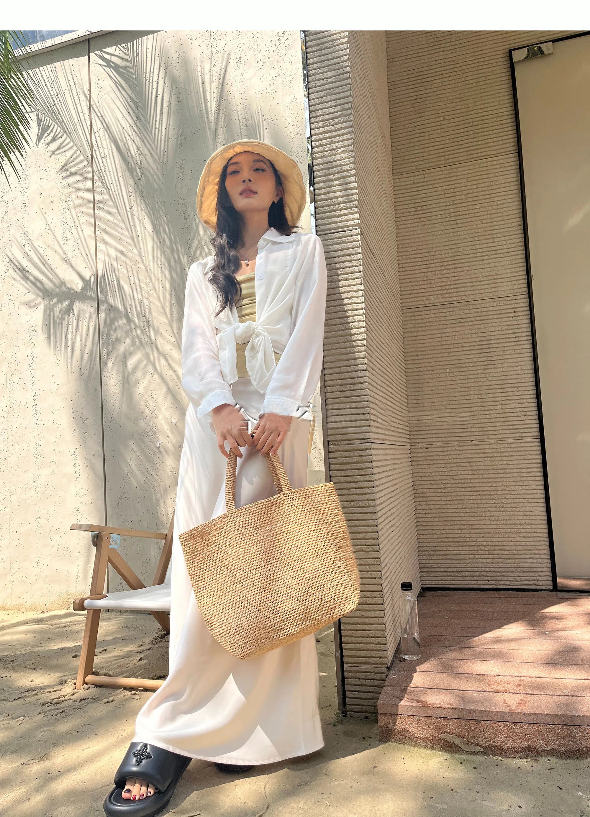 Woman in a white outfit with a straw hat and bag standing outdoors.