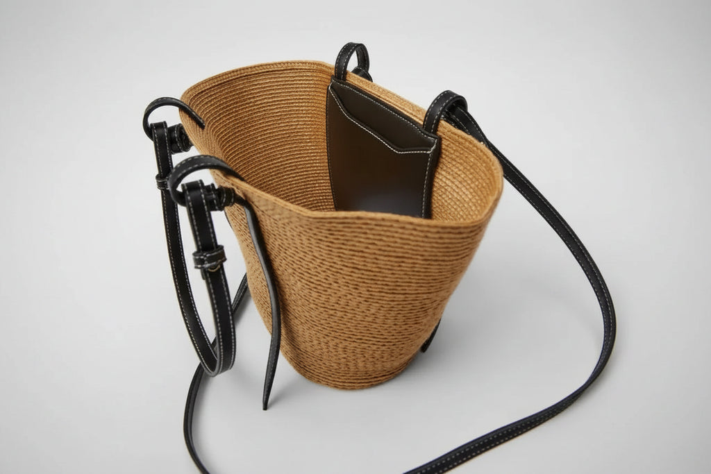 A straw tote bag by Yue Jabon, featuring a natural tan woven body, black leather handles, a black interior lining, and a long black shoulder strap, shown on a light gray background.