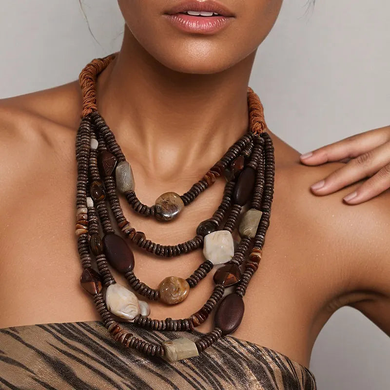 A close-up of a model wearing a multi-strand statement necklace made of dark brown beads, woven cords, and polished natural stones in earthy brown, beige, and gray tones.