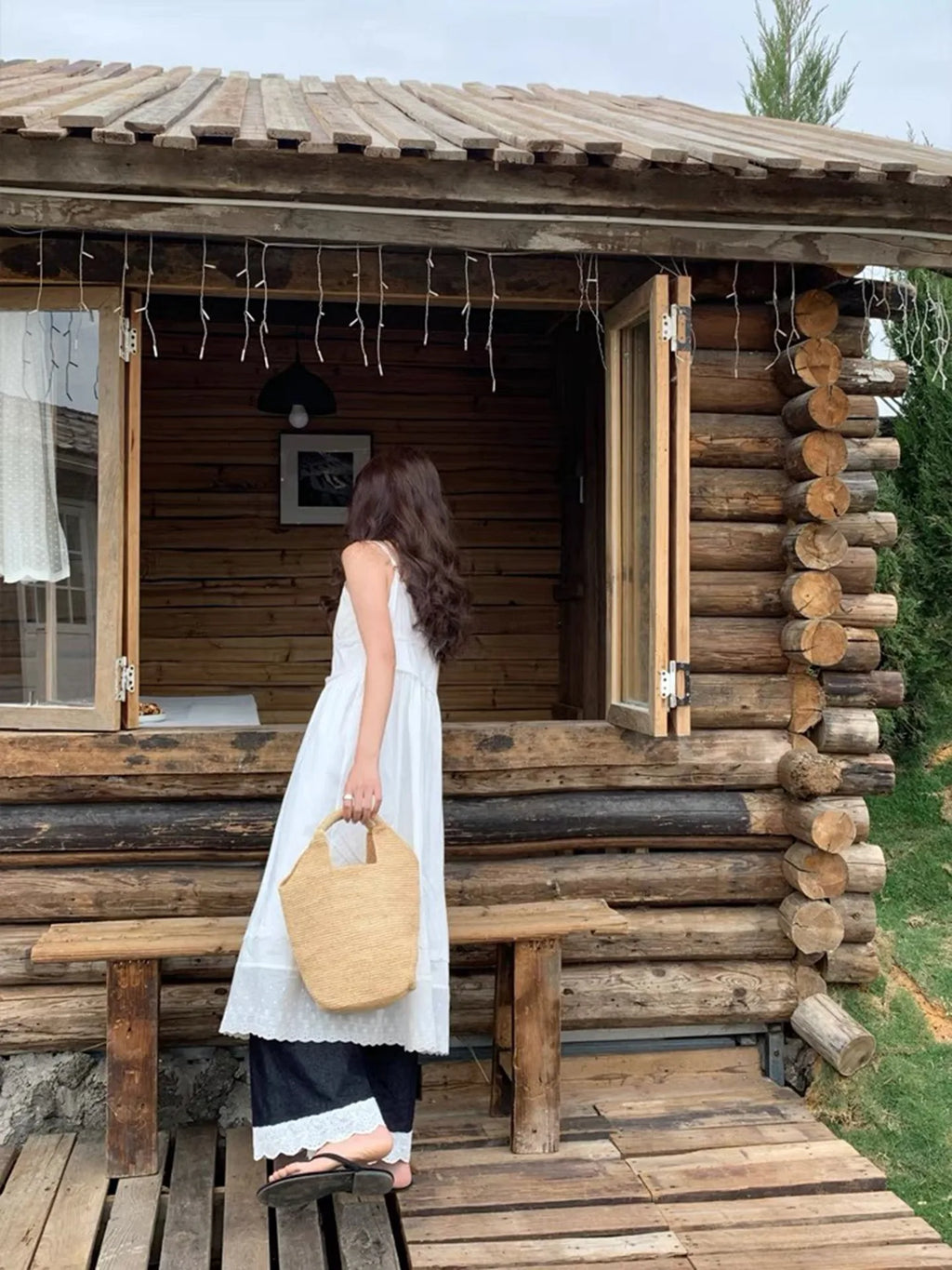 Woman in a white dress standing in front of a wooden cabin holding a beige bag.