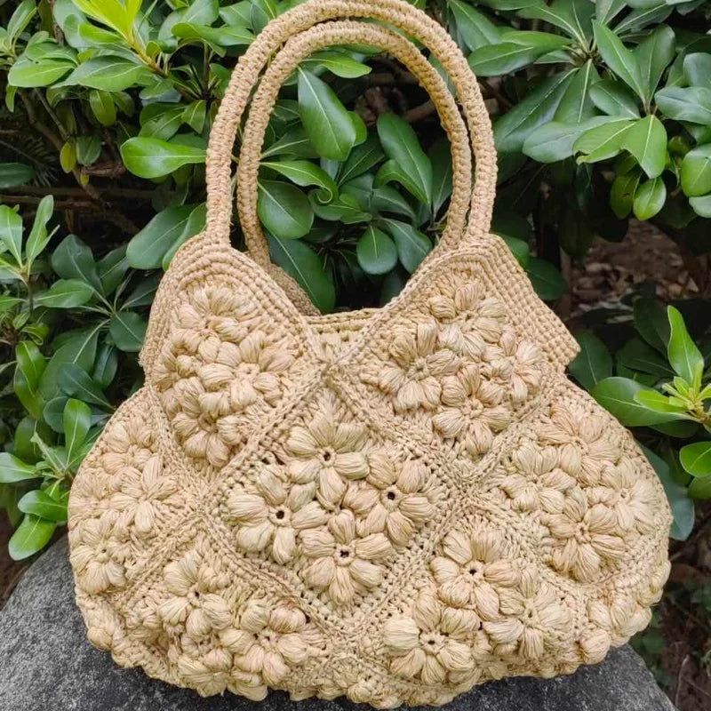 A natural beige woven straw handbag with textured crochet floral patterns and two top handles, resting on a stone surface in front of green foliage.