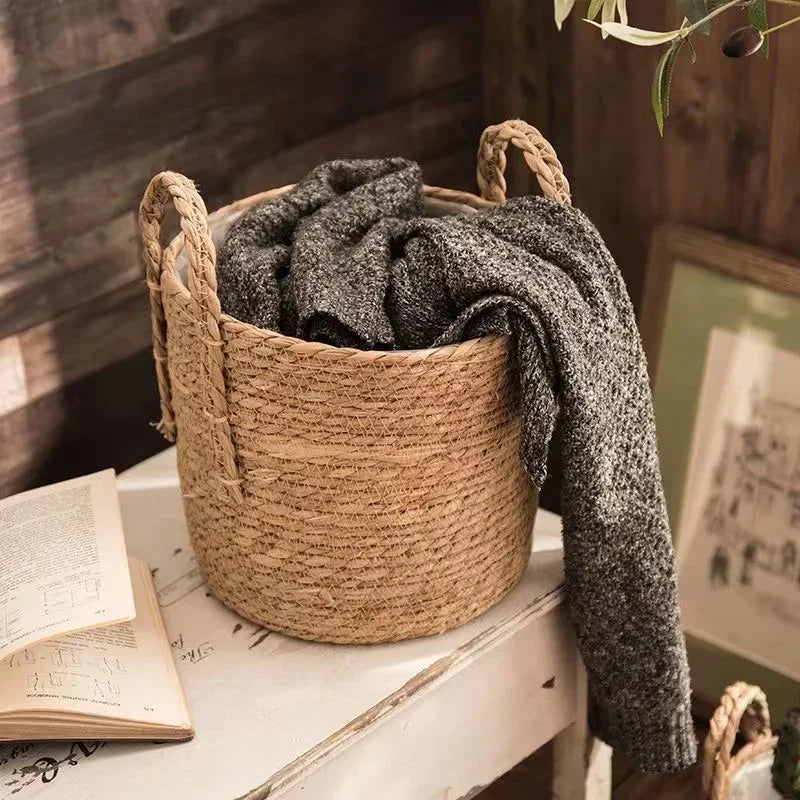 Woven basket with a gray sweater on a wooden surface with books and a framed picture in the background.