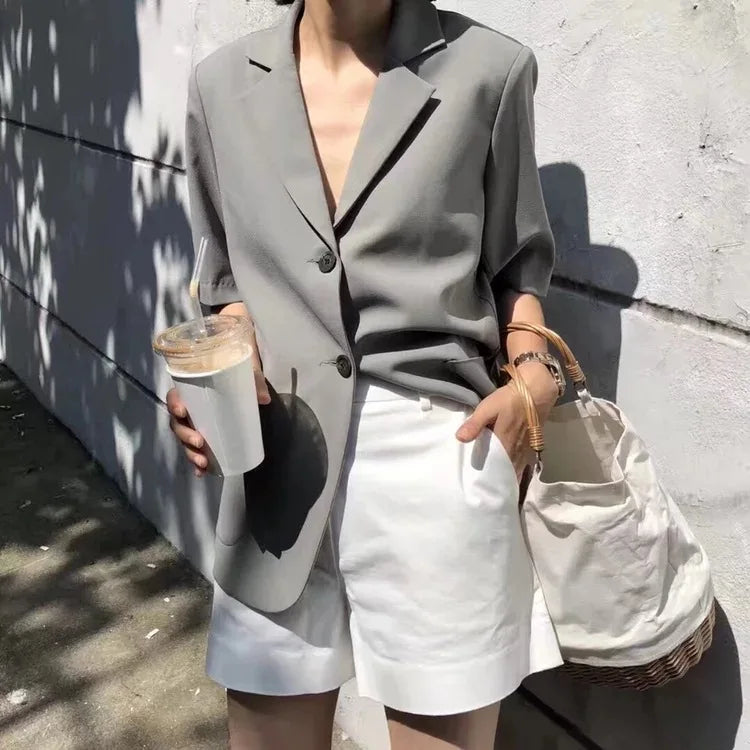 Person wearing a gray blazer and white shorts, holding a drink and a bag, against a concrete wall.