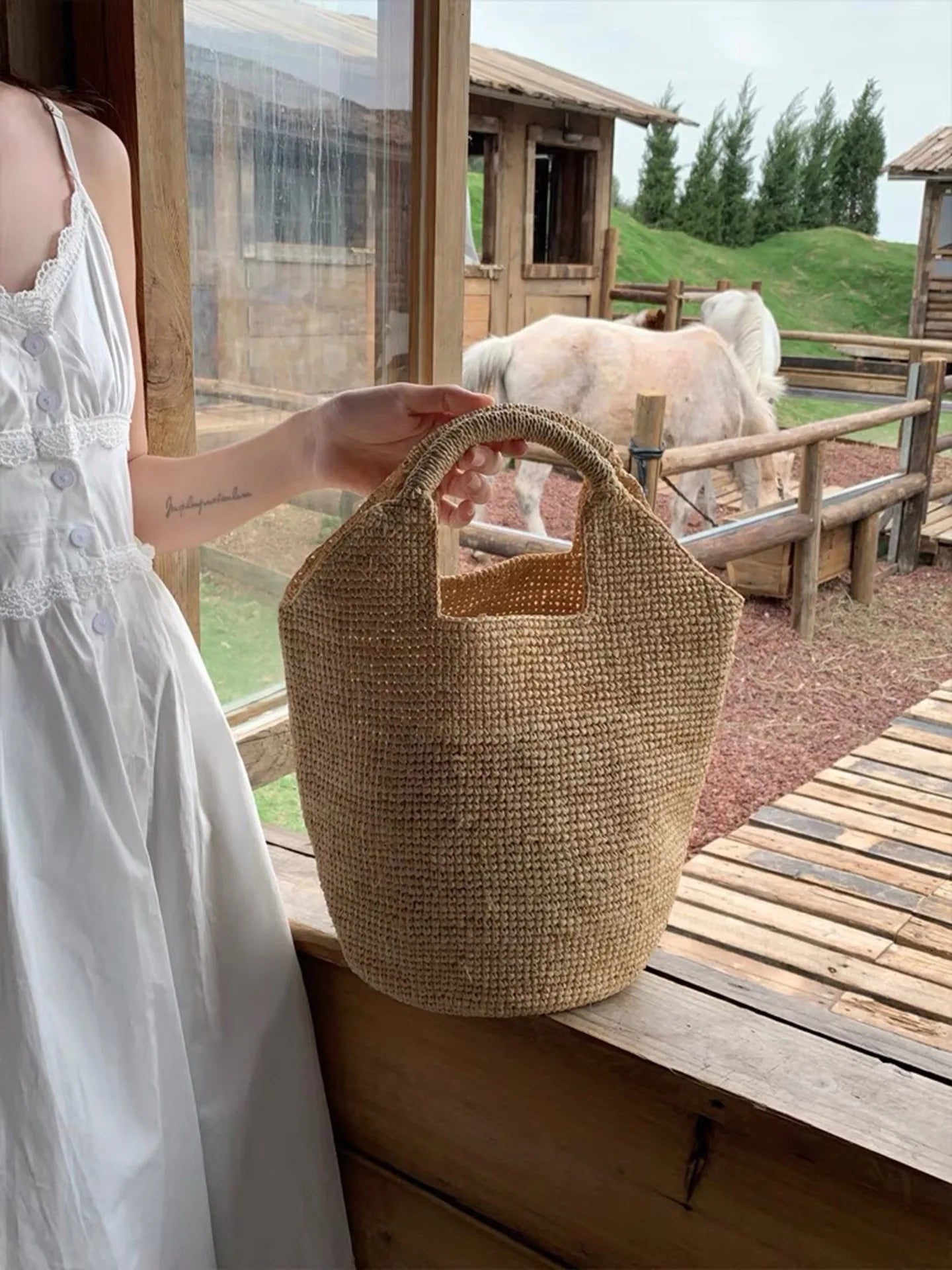 Person holding a woven bag with a scenic background of animals and wooden structures.
