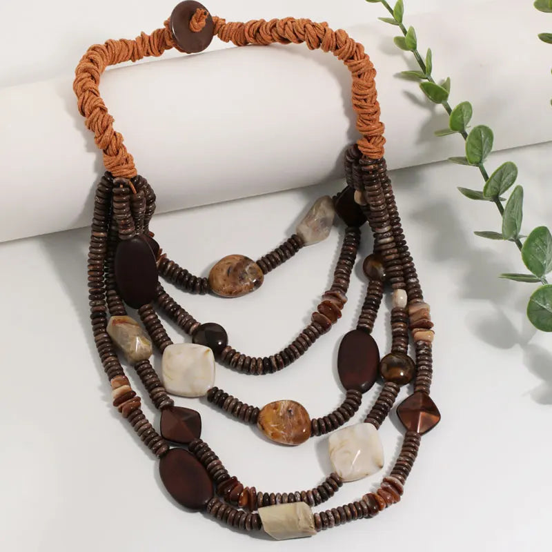 A multi-strand statement necklace featuring dark brown beaded strands, polished brown and cream stones, and a braided orange cord collar, displayed on a white surface with a green plant sprig.