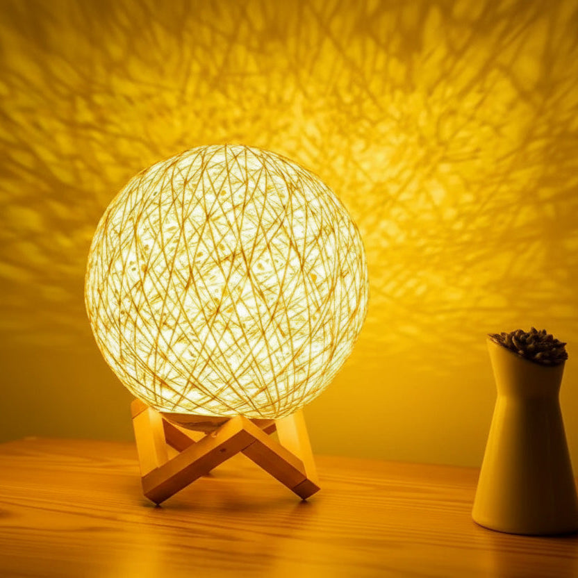 Woven ball lamp on a wooden stand with a soft glow against a warm yellow background.