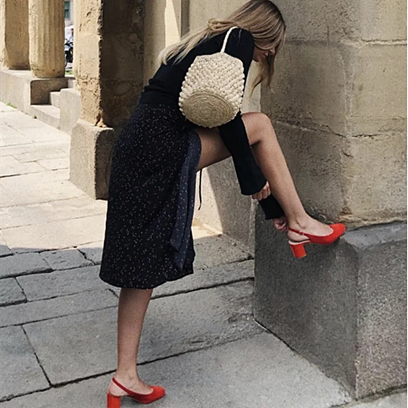 Woman sitting on a stone step wearing a black dress, red shoes, and a straw bag.